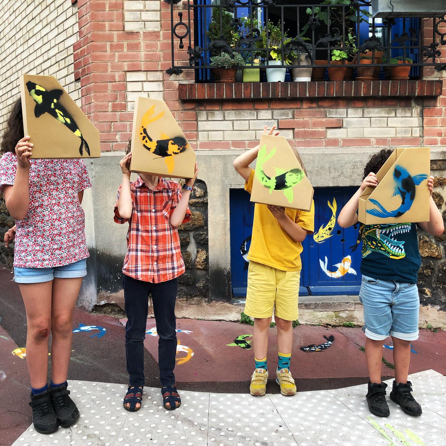 Quatre participants présentent leurs carpes koï peintes au pochoir devant une façade ornée de poissons